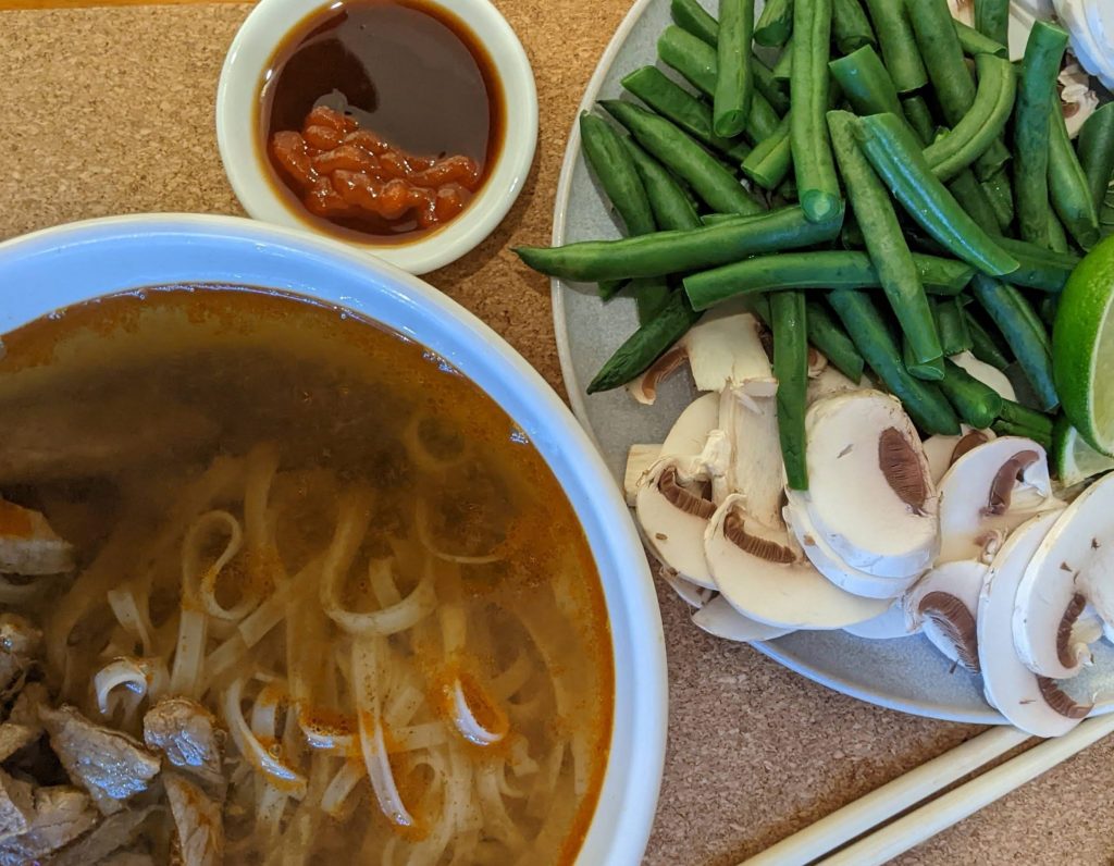 A bowl of Mr Chen's beef pho with a plate of green beans and mushrooms and a sauce dish with gluten free hoisin and sriracha.