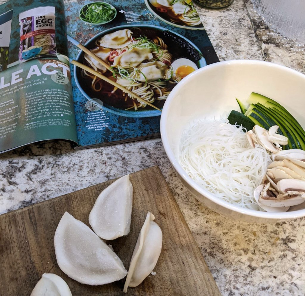 An open magazine with the recipe, a bowl with cooked noodles, zucchini slices and mushrooms. Frozen dumplings sitting on a chopping board.
