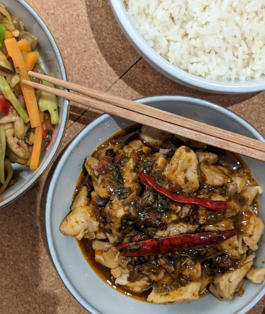 Three bowls, one off screen with mixed vegetables. One with cooked white rice. Middle bowl has Mr Chen's Thai basil chilli, two large dried chillis sit on top of the cooked chicken.