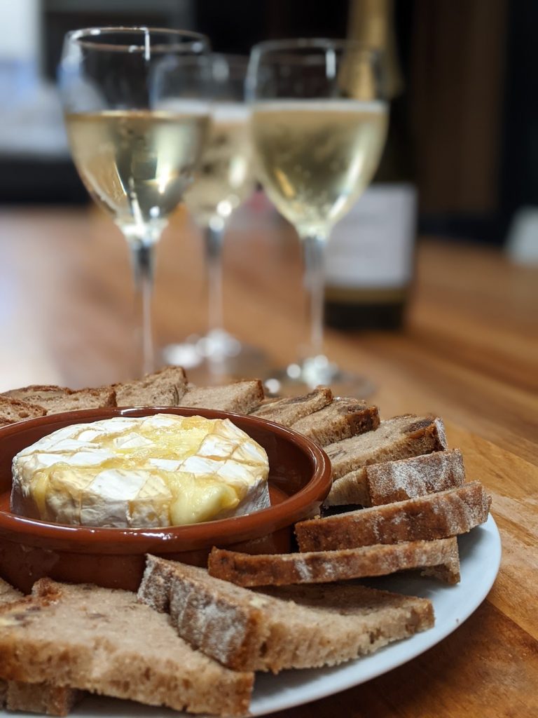 A dish with oozing baked camembert, surrounded by sliced bread with three glasses of champagne in the background.