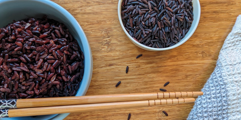 Cooked black rice in a bowl with chopsticks. A measuring scoop of raw black rice.