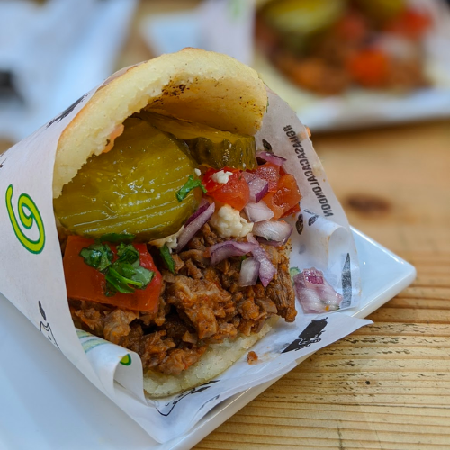 An arepa with a wide opening showing meat, pickles, tomato and red onion.