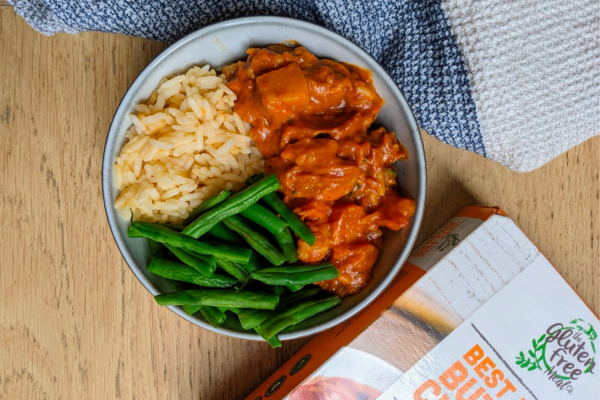 A bowl of rice, green beans and butter chicken. A box for gluten free meal co is off to the side and a blue and white cloth is wrapped around the bowl.