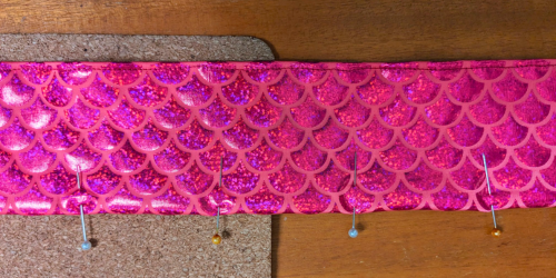 A strip of pink scaled fabric. One side is stitched with a straight stitch, the other is folded and held with pins.