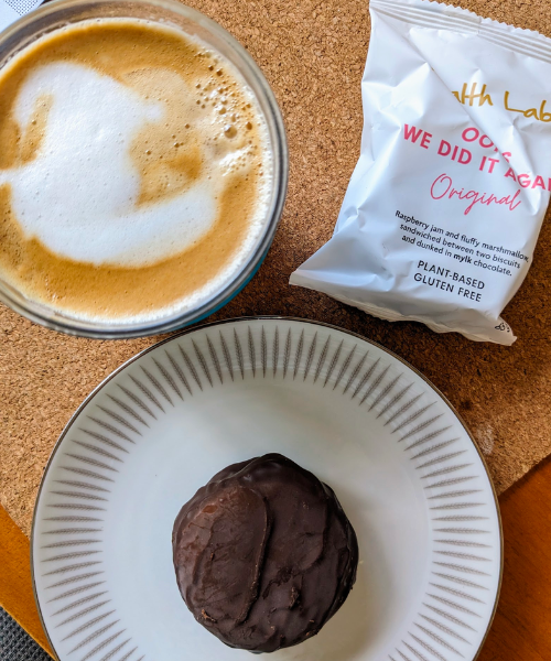 Top view image of a cup of coffee, a chocolate cookie on a plate and a packet of oops we did it again. 