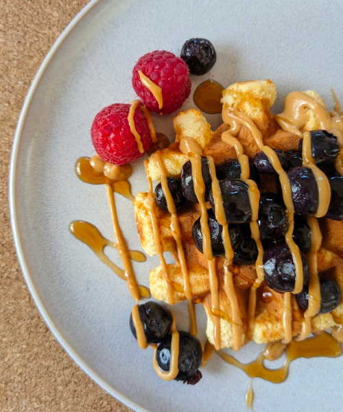 A waffle on a plate topped with blueberries, drizzled with peanut butter and maple syrup. Two raspberries to garnish.