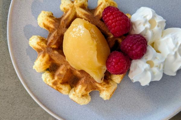 A waffle on a plate with a scoop of lemon curd, raspberries and whipped cream.
