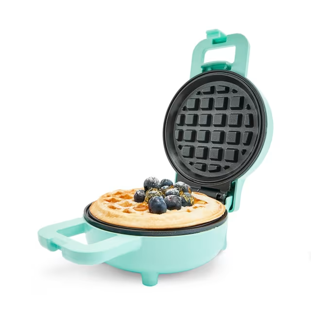 A teal waffle iron with a waffle inside topped with blueberries.