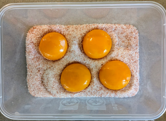 A takeaway container half filled with pink salt, four egg yolks sit inside.