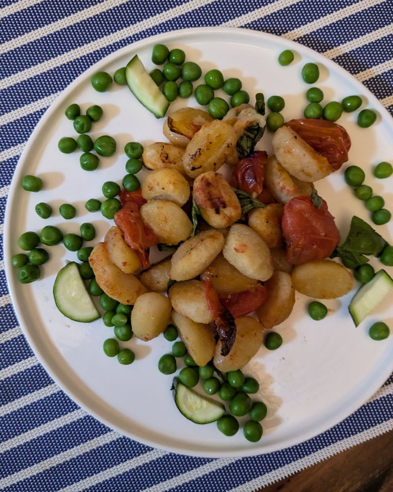 A plate of gnocchi and roasted tomato surrounded by peas and zucchini pieces.