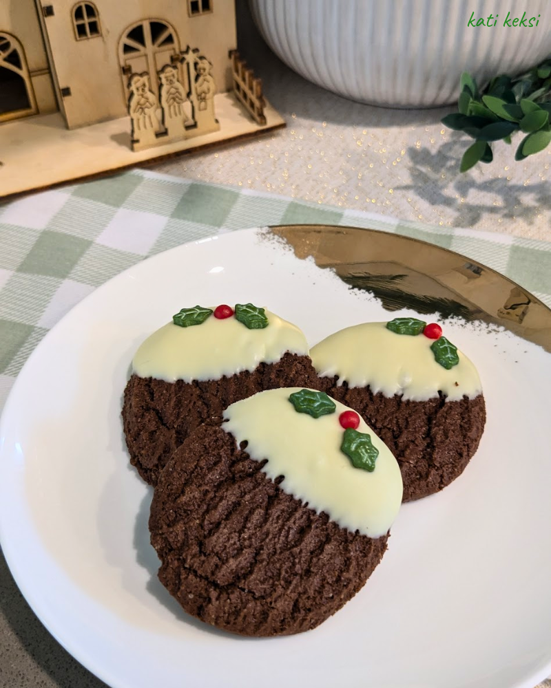 Three cookies decorated to look like Christmas pudding on a white plate with a green gingham cloth in the background.