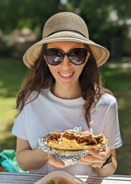 A woman in a white shirt, sunglasses and hat holding a large arepa.